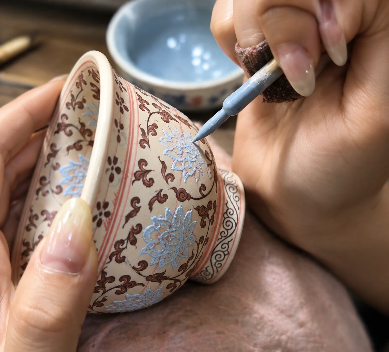 An artisan hand-painting a lotus motif onto a porcelain teacup with a fine brush, filling the petals in pale blue