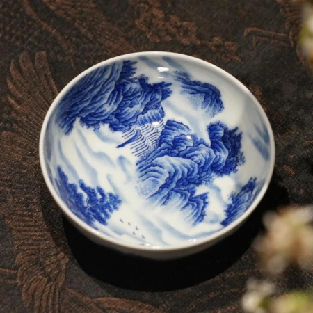 A hand-painted blue and white porcelain dish with a sweeping mountain landscape motif, on a dark brocade cloth