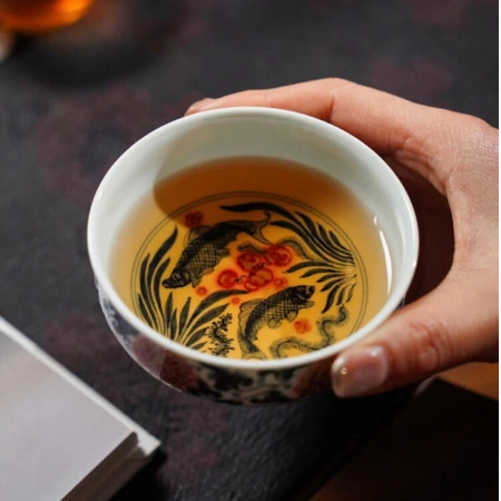 A hand holding a porcelain teacup of tea, its interior painted with a pair of fish and waterweed