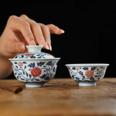 A hand lifting the lid of a hand-painted blue and white gaiwan with iron-red floral scrolls, beside a second gaiwan
