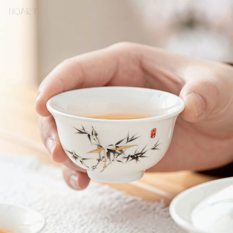 One of the tea cups from the lucky bamboo gaiwan tea set, held between finger and thumb — black leaves, gold stems, red seal.