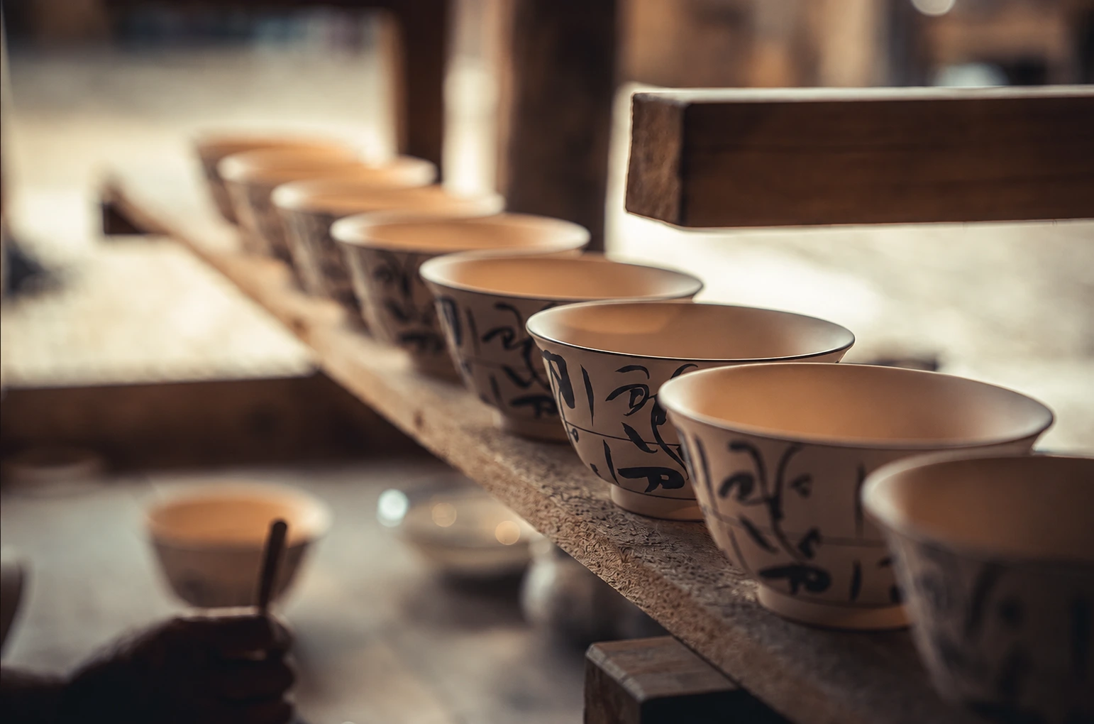A row of hand-painted porcelain cups with floral scroll designs lined up on a wooden shelf in a pottery studio