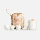 A porcelain tea set on plain white: a brewing cup with its rose-topped lid and the strainer that nests inside it, three tea cups and a glass cup, in front of the floral carrying case.