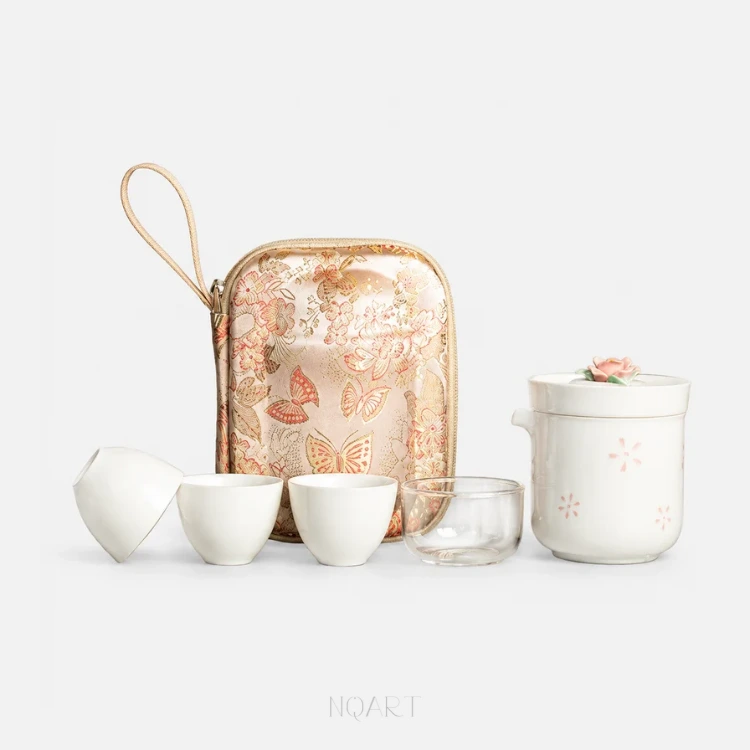 A porcelain tea set on plain white: a brewing cup with its rose-topped lid and the strainer that nests inside it, three tea cups and a glass cup, in front of the floral carrying case.