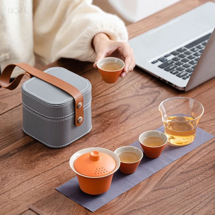 Seen at an angle, this portable tea set with case has its leather handle standing up, the gaiwan, three cups and the glass pitcher set out beside it.