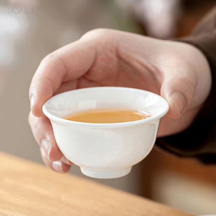 Cupped in one hand, one of the small tea cups holds a shallow pour — thin white porcelain, flared at the lip and set on a small foot.