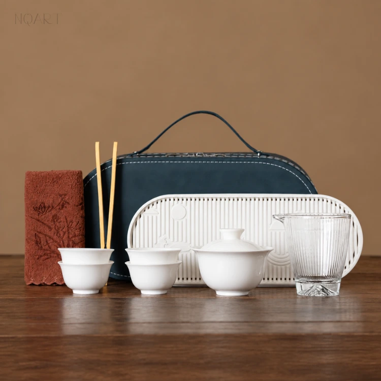 Low light on the wooden table, the tea ceremony travel set stands with its handle up — navy case, slotted tea tray and white porcelain arranged along one line.