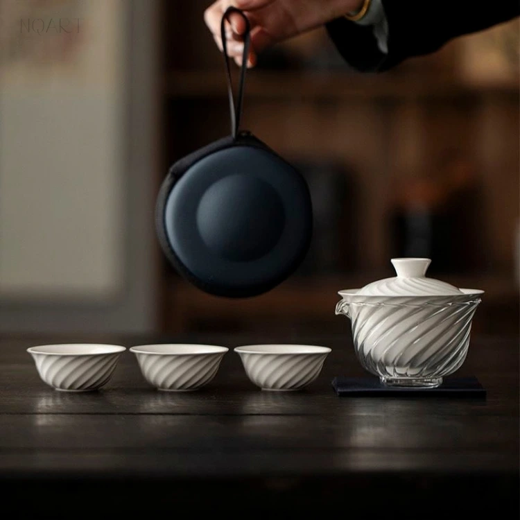 Dangling from its strap, this tea set for hiking hangs clear of the table above three cups and the glass pitcher it is about to be unpacked onto.
