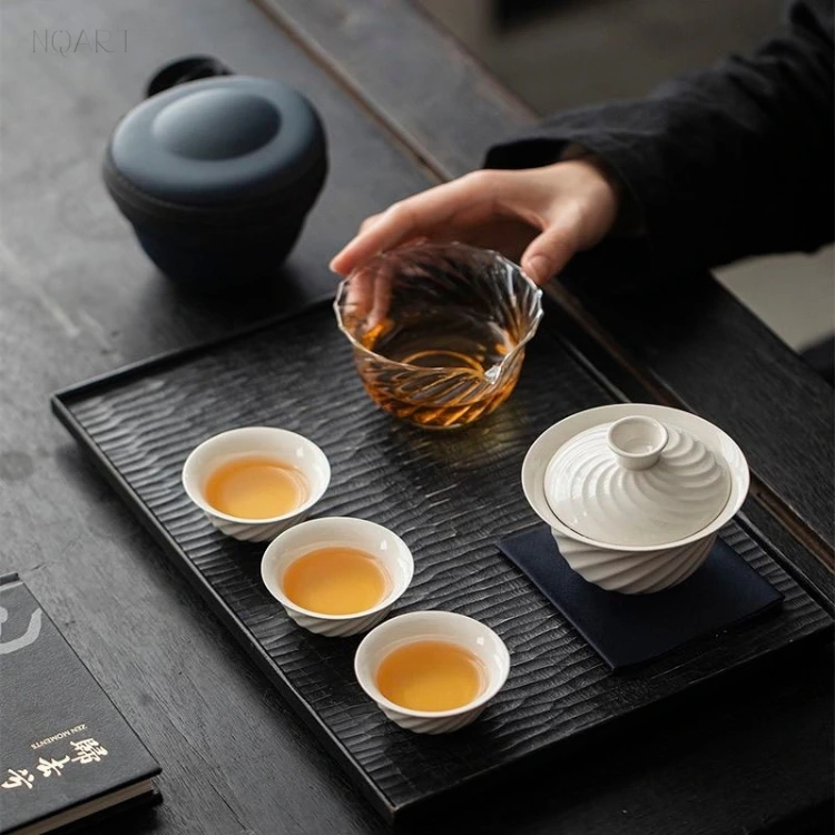 On the black mat, this tea set for outdoor use is spread across the corner of a table — gaiwan on its cloth, three cups, and the glass pitcher under a reaching hand.