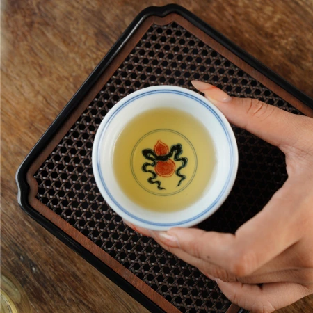 A hand holding a porcelain teacup above a wooden tea tray, its interior painted with a dark branch bearing red fruit
