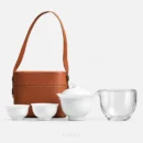 A four-piece travel tea set with bag, laid out in a row: white porcelain gaiwan, two cups and a glass pitcher, the tan leather bucket bag standing behind them.