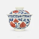 Lid on, this gaiwan reads as one rounded form — blooms of lotus and peony in underglaze red set among a scrolling blue vine that carries from the bowl up onto the lid.
