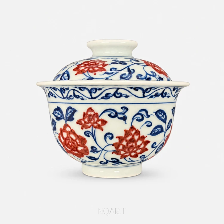 Lid on, this gaiwan reads as one rounded form — blooms of lotus and peony in underglaze red set among a scrolling blue vine that carries from the bowl up onto the lid.