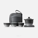 Set straight on, against white: a spouted gaiwan tea set — the bowl with its pouring lip and pierced lid, three two-tone cups, and the black hard case standing behind them.